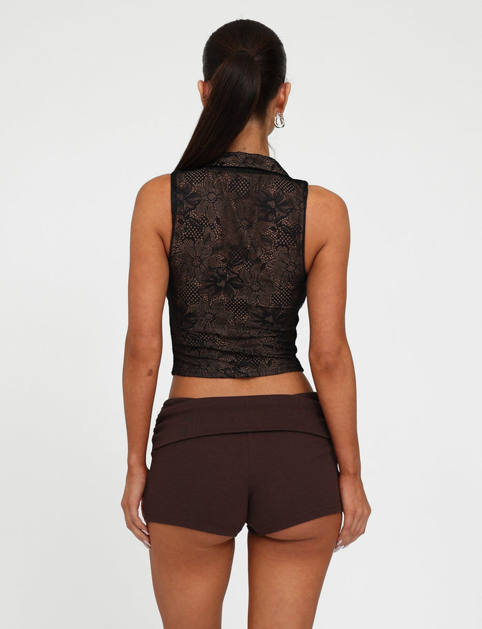A woman with a high ponytail stands with her back to the camera, donning the ALDINA TOP - BLACK : LACE by TIGERMIST and brown high-waisted shorts. She accessorizes with hoop earrings and is set against a plain white background.