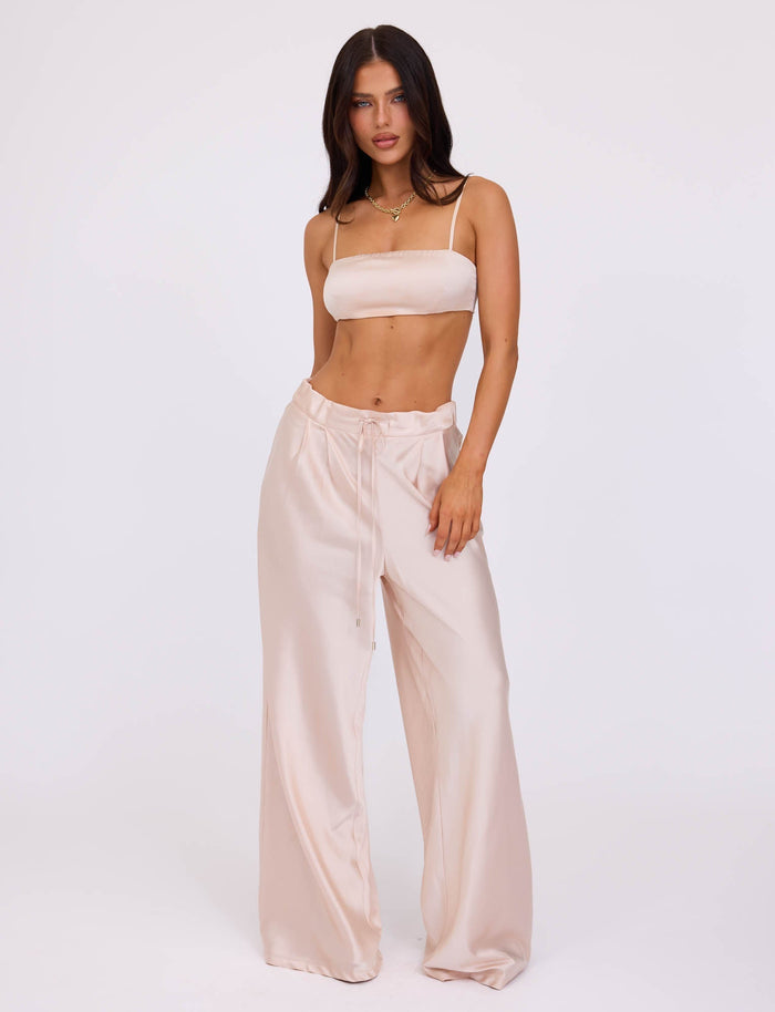 A woman with long dark hair stands against a plain white background, wearing the matching champagne-colored satin crop top and ALINA PANT - WHITE : IVORY from TIGERMIST. She gazes directly at the camera, one hand resting in her pocket, and the other hanging by her side.