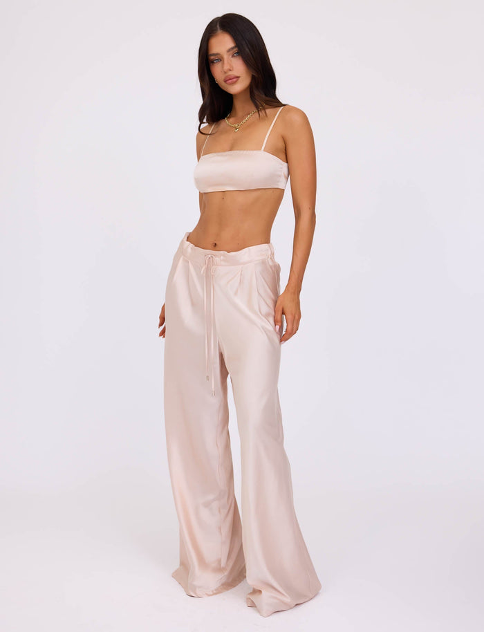 A woman stands against a plain backdrop, wearing the ALINA PANT - WHITE : IVORY from TIGERMIST, paired with a light beige crop top. She has long dark hair and is posing with one hand on her hip while looking directly at the camera.
