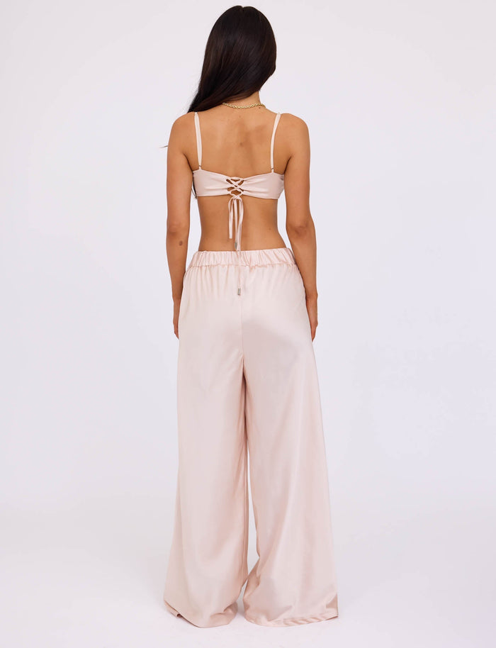 A woman with long dark hair is facing away from the camera, wearing a matching light beige satin crop top with thin straps and back ties, paired with the ALINA PANT - WHITE : IVORY high-waisted wide-leg satin pants from TIGERMIST. The background is plain white.
