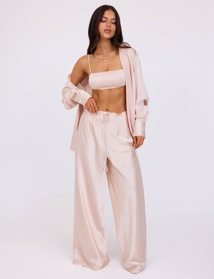 A person with long brown hair stands wearing a light pink silk pajama set from TIGERMIST, featuring a cropped camisole and wide-legged pants, paired with the ALINA SHIRT - WHITE : IVORY draped unbuttoned over their shoulders. They pose with one hand on their hip against a plain white background.