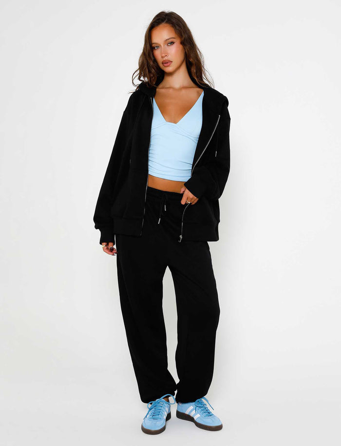A woman stands against a plain white background, donning a black zip-up hoodie from TIGERMIST, ALISH PANT in black from TIGERMIST, a light blue crop top, and blue sneakers. She has long, wavy brown hair and adopts a relaxed pose, with one hand in her pocket and the other holding the bottom of her hoodie.