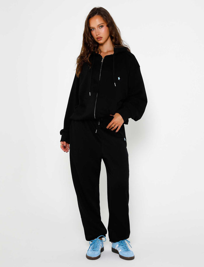 A woman with long, wavy hair wears the ALISH PANT in black by TIGERMIST, a black zip-up hoodie over a black top, and light blue sneakers. She stands against a plain white background with one hand in her pocket and a relaxed expression.