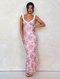 A woman with dark hair up poses confidently against a light textured wall, wearing the TIGERMIST AMARAN DRESS - PINK : CAMO FLORAL with white straps and neckline, paired with white heeled sandals.