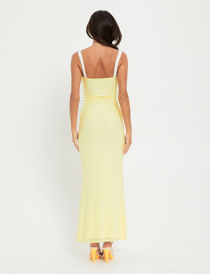 A woman with long dark hair is standing with her back to the camera, wearing an AMARAN DRESS in yellow (Lemon) from TIGERMIST. She has orange high-heeled shoes and is standing against a plain white background.