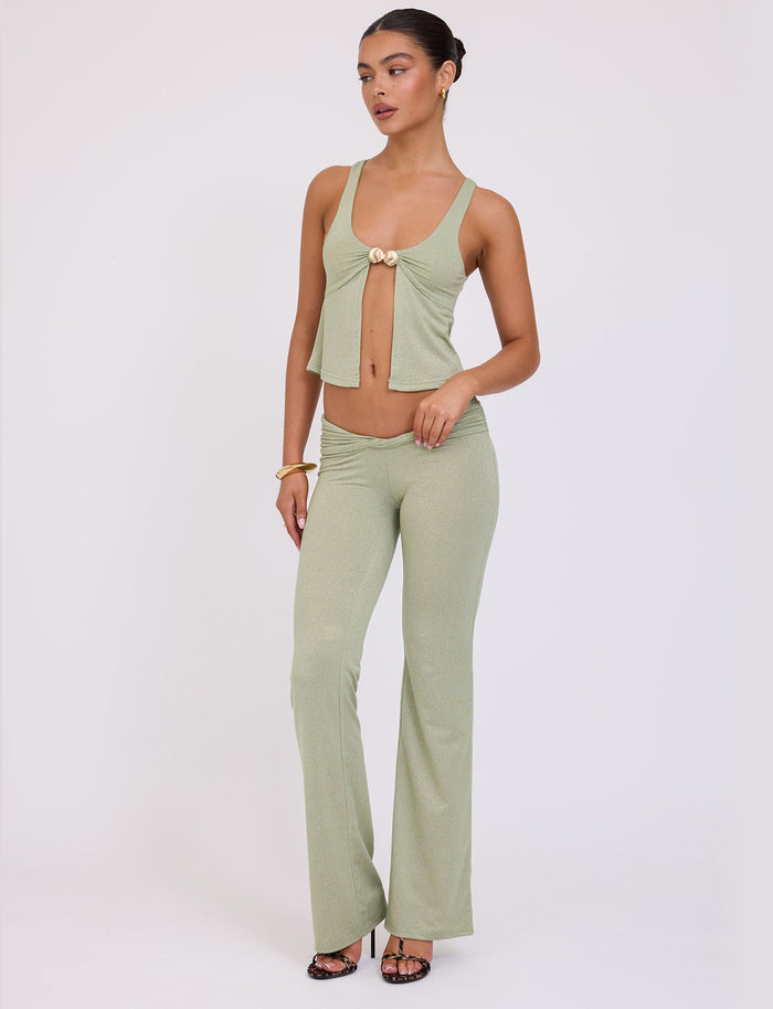 A woman stands posing against a plain white background, showcasing a chic Tiger Mist ensemble. She is wearing the Amethyst Pant in Green Sparkle, paired with a coordinating light green, sleeveless top featuring a front tie. She accessorizes with gold bracelets and black strappy heels. Her hair is neatly styled in an updo.