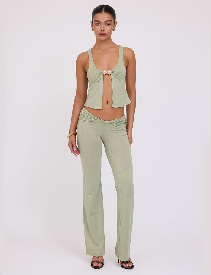 A person stands against a white backdrop wearing the LIGHT GREEN SPARKLE "AMETHYST" SLEEVELESS TOP by TIGERMIST, featuring a front tie at the bust, paired with matching high-waisted flared pants. They accessorize with hoop earrings, a bracelet, and open-toe heels. Their expression is neutral and their hairstyle is sleek.