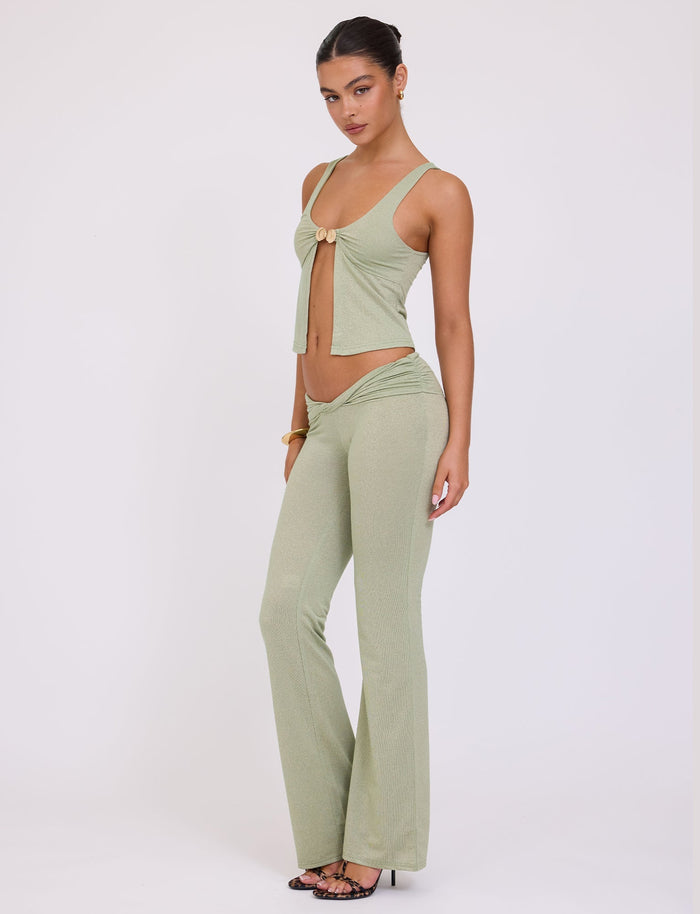 A woman stands against a plain white background, wearing the chic green sparkle Amethyst Top by TigerMist. Her stylish ensemble includes a sleeveless top with a front tie and high-waisted, flared pants. With her hair pulled back and black strappy heels, she effortlessly exudes chic elegance.