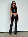 A woman with long dark hair wears a black sleeveless crop top and the ANEYA PANT - BLACK CORD, which features a low-rise waist and large square buckles. The flared silhouette accentuates her figure as she stands confidently in black sunglasses and open-toe heels against a plain white background.