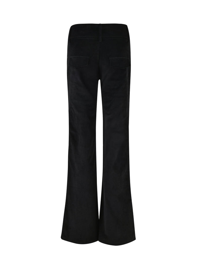 A woman with long dark hair wears a black sleeveless crop top and the ANEYA PANT - BLACK CORD, which features a low-rise waist and large square buckles. The flared silhouette accentuates her figure as she stands confidently in black sunglasses and open-toe heels against a plain white background.