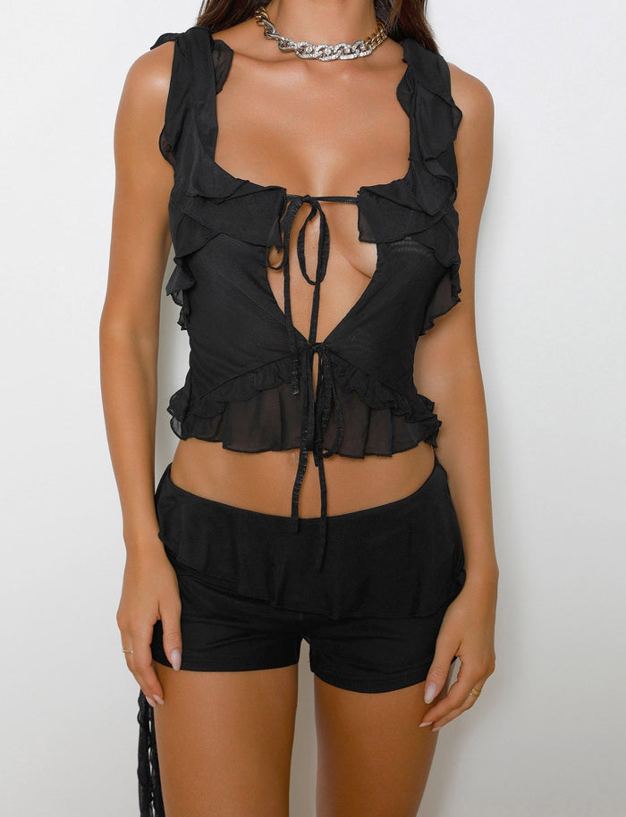 A person wearing the ANISHA TOP - BLACK from TIGERMIST, a sleeveless ruffled crop top with a tie-front, paired with matching black Tiger Mist shorts is standing against a plain white background. The outfit includes a chain necklace, and the person's hands are hanging naturally at their sides.