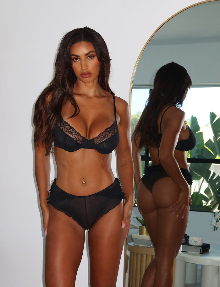 A woman with long, wavy hair stands in front of a large mirror. She is wearing the ARA SET - BLACK lingerie from TIGERMIST and has a belly button piercing. The background reveals a well-lit room with a plant near the window.