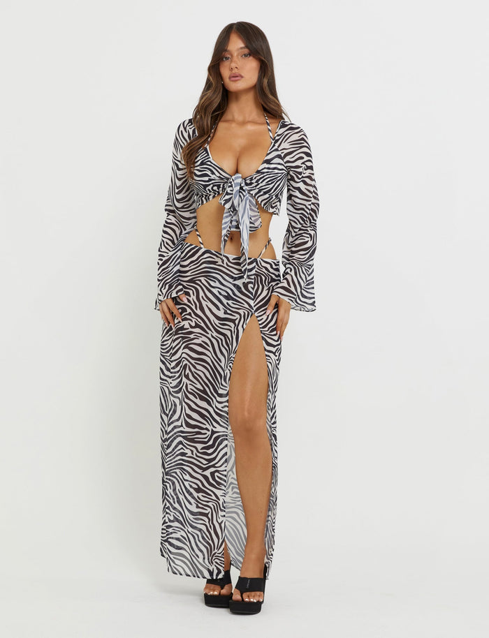 A woman stands against a plain white background wearing the TIGERMIST AYESHA SKIRT in black zebra print. The ensemble includes a long-sleeve, tie-front crop top paired with a high-slit maxi skirt. She has long hair, wears black platform heels, and exudes confidence.