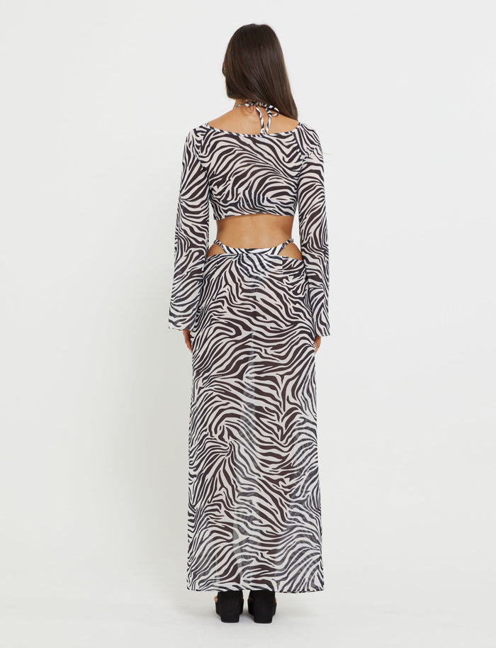 A person with long dark hair is seen from the back wearing a stunning TigerMist Ayesha Skirt in zebra print paired with a long-sleeved top featuring cutout details at the sides and back. The skirt reaches their ankles, and they are wearing black heeled shoes. The background is plain white.