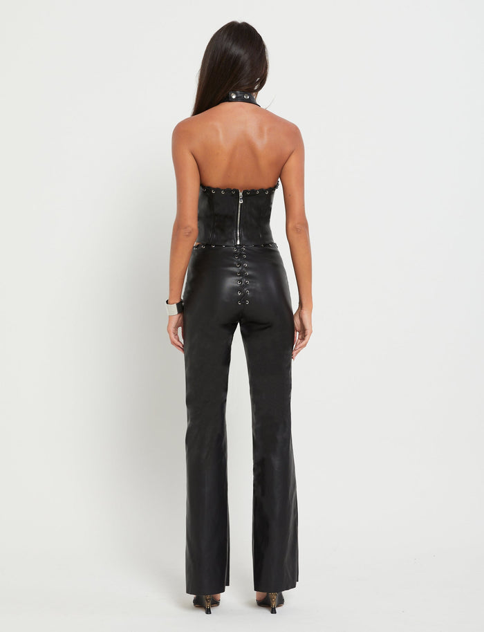 A woman with long, straight hair is facing away from the camera, showcasing her stylish TIGERMIST outfit. She is wearing a black leather halter-top jumpsuit with lace-up details on the back, paired with high heels. The crisp white background accentuates her chic BOSCO PANT - BLACK: BLACK PU attire.