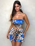 A woman with long dark hair wears a stylish TIGERMIST BRIXLEY TOP - MULTI : LEOPARD, featuring a blue and leopard print pattern with floral designs. The top has a unique cutout at the waist, and she pairs it with a matching skirt that has a flowy A-line shape. She stands against a light-colored background.