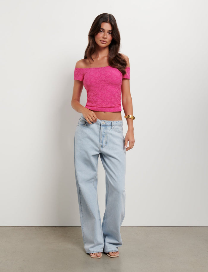 A woman stands confidently in front of a white background, embodying TIGERMIST style with the CAIDEN TOP in hot pink lace and light blue high-waisted wide-legged jeans. She is accessorized with a gold bracelet and strappy heels, her dark hair styled in loose waves.