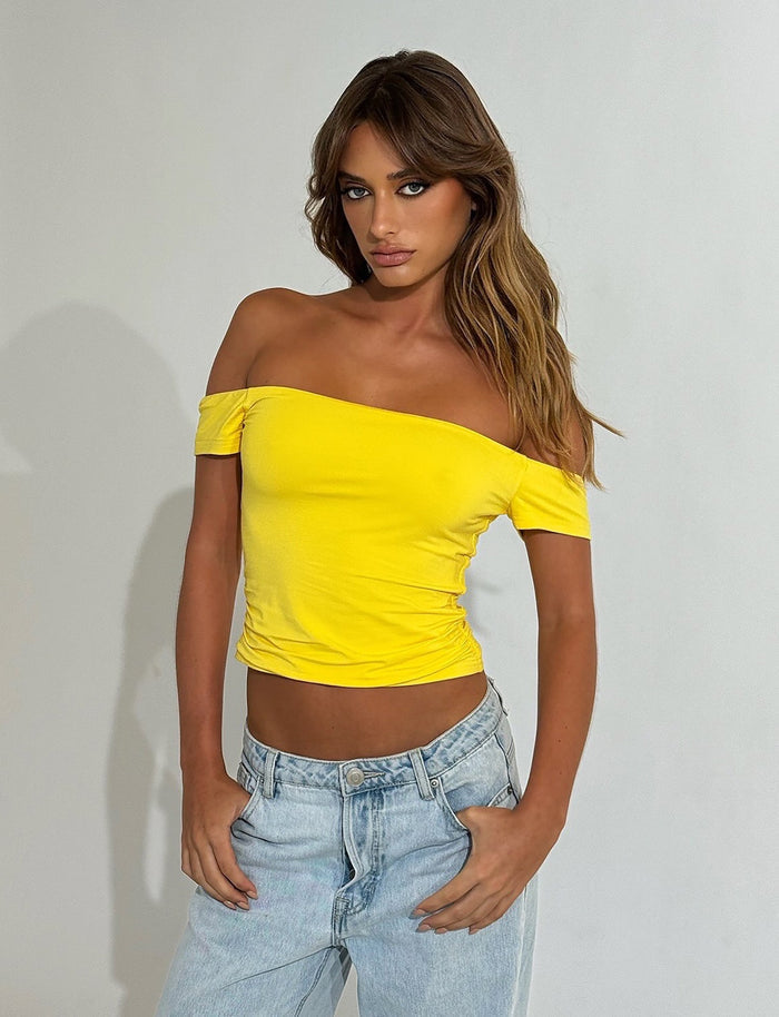 A person with long, wavy brown hair stands against a plain white background, wearing the bright yellow CAIDEN TOP from TIGERMIST and light blue jeans. They have one hand resting on their hip and a serious expression on their face.