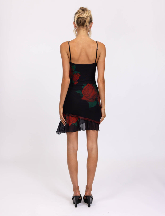 A person with blonde hair styled in an updo is standing facing away from the camera, wearing the TIGERMIST CARLY DRESS - BLACK : MACRO ROSE- RED ROSE. This sleeveless dress features thin straps and a red rose pattern, with a slight ruffle at the hem. They complete the look with black high heels.