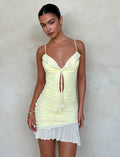 A woman wearing the CARLY DRESS - YELLOW : LEMON by TIGERMIST, a light yellow ruched mini dress with shoulder straps and a front tie, stands against a simple beige background. Her hair is pulled back, and she is accessorized with gold earrings and bracelets. She has a neutral expression with her hands by her sides.