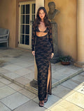 A woman stands outside on a tiled patio in front of a door and near a potted plant. She is wearing the CHRISTY LACE L/S MAXI DRESS - BLACK : LACE from TIGERMIST, featuring a high slit and cutout detail at the midriff. She has long, wavy hair and is looking directly at the camera.