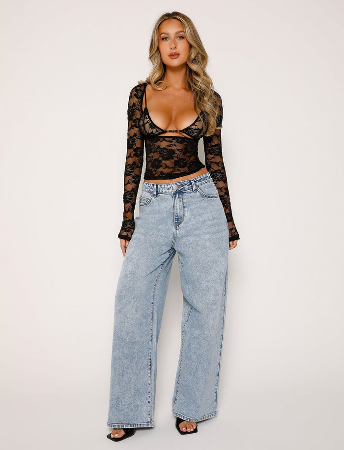 A woman stands against a plain background wearing the CHRISTY LONG SLEEVE TOP - BLACK : LACE from TIGERMIST and high-waisted, light-wash baggy jeans. She has long, wavy blonde hair and is accessorized with black open-toe heels. Her outfit seamlessly blends edgy and casual styles.