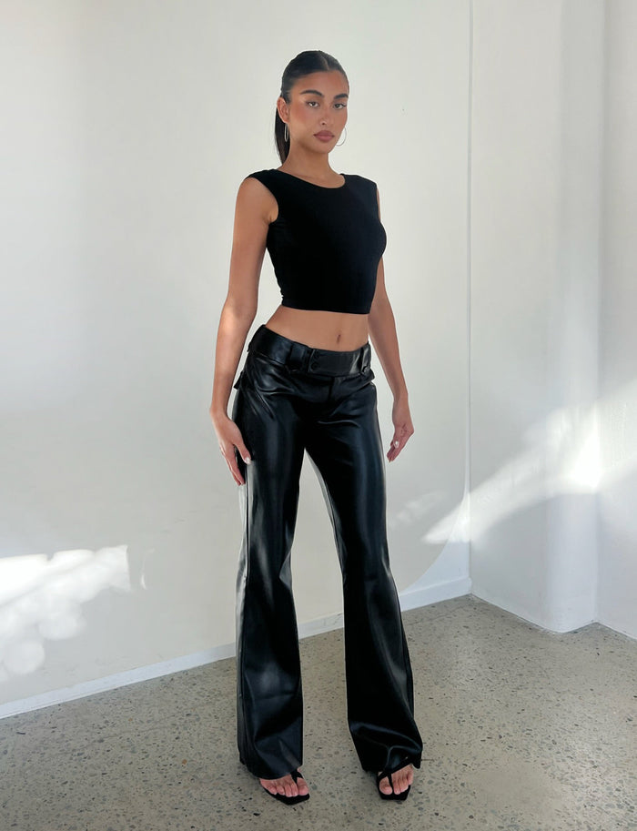 A person with long hair in a ponytail is posing in a brightly lit, minimalistic space. They're wearing the TIGERMIST CHRISTY TOP - BLACK, high-waisted black leather pants, and black open-toe shoes. Their expression is neutral and their hands are by their sides.