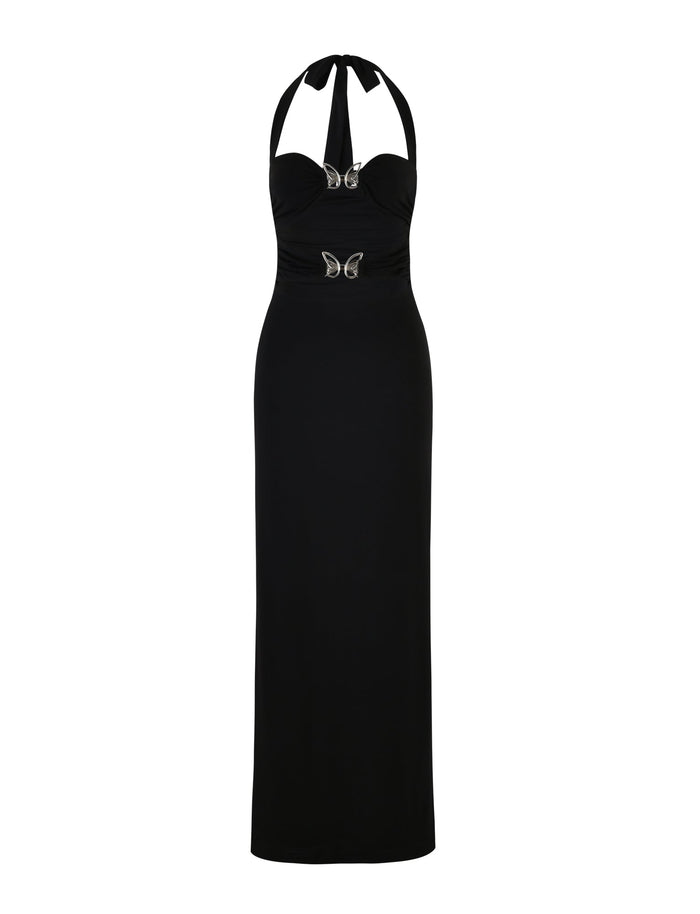 A long, elegant black halter-neck dress called the CORALINE DRESS - BLACK from TIGERMIST with a fitted silhouette. The bodice features ruching and two decorative butterfly-shaped embellishments, creating a glamorous and sophisticated look.