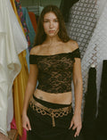 A woman stands confidently in front of a backdrop of various fabrics. She is wearing the CYD TOP - BLACK, an off-the-shoulder lace top from TIGERMIST, paired with a gold chain belt with circular links and a black skirt. Her long dark hair cascades down, revealing a small tattoo on her right upper chest.