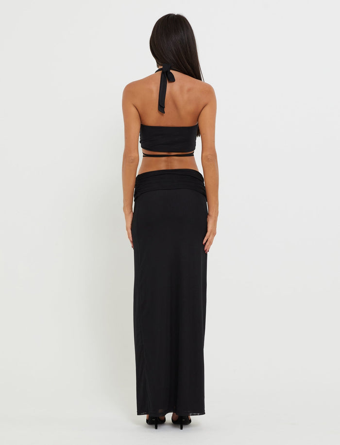 A woman with long dark hair stands facing away, wearing a chic black halter top and the flowing TIGERMIST Cynthia Maxi Skirt in black. The background is plain white.