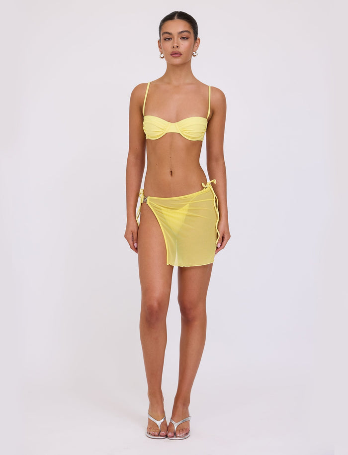 A woman stands against a plain white background, wearing a yellow TIGERMIST bikini top with thin straps and the CYNTHIA MINI MESH SKIRT in pale yellow, tied at the waist to reveal matching bikini bottoms. She has her hair tied back, accessorizes with earrings, and wears clear high-heeled sandals.