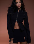 A woman with long, wavy dark hair is wearing the DAMIRA JACKET - BLACK and a matching skirt with a small slit from TIGERMIST. She has a neutral expression and is set against a plain brown background.