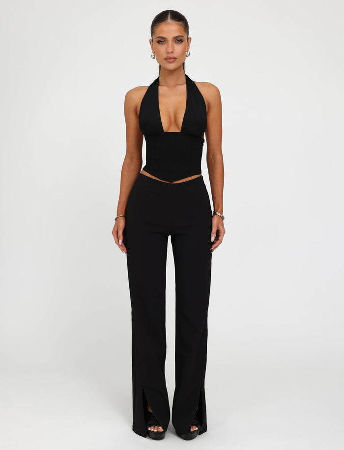A woman stands against a plain white background. Sporting a sleek outfit from TIGERMIST, she wears a black halter top with a deep V-neckline and the DAMIRA PANT in black, which features high-waisted design and slits at the front hems. Her hair is pulled back, accessorized with small hoop earrings and a bracelet.