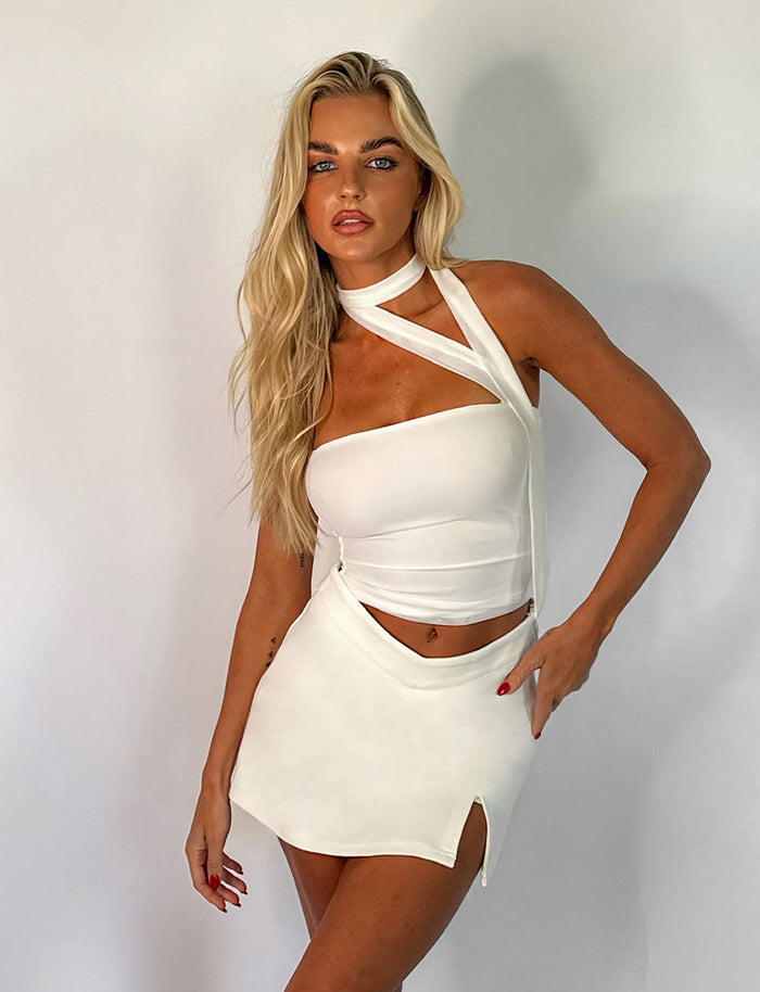 A woman with long blonde hair poses against a white background, wearing a white asymmetrical sleeveless top paired with the DAPHNIE SKORT - WHITE from TIGERMIST. The mini-length skort highlights her red nails as she gazes confidently at the camera.