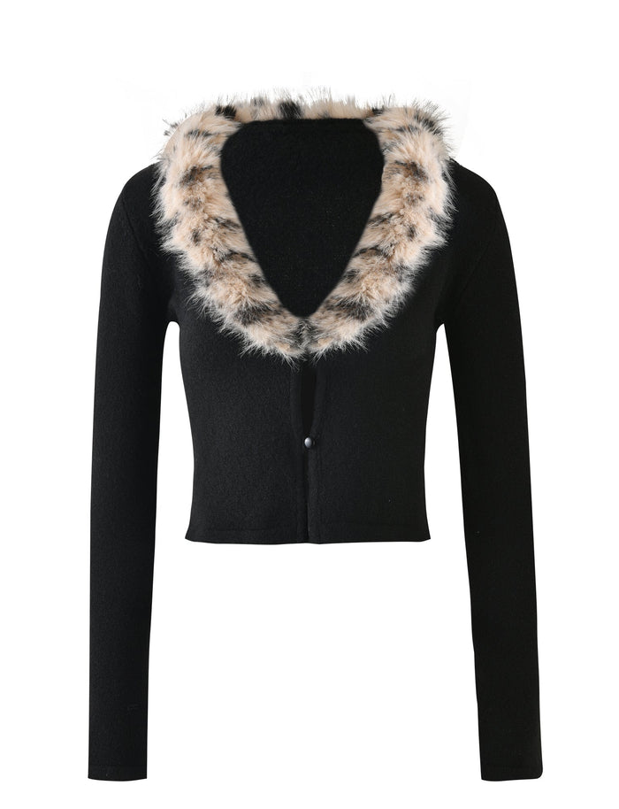 The DARJA CARDIGAN - BLACK from TIGERMIST is a black long-sleeved crop top featuring a deep V-neck collar. It is adorned with a thick, fluffy faux fur trim in a white and black spotted pattern and has a single button closure at the front.