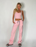 A woman with long blonde hair, clad in a light pink tank top and matching Tigermist Darja Pant in baby pink, poses confidently against a plain white background. With her hands in her pockets and pink and grey sneakers on, she exudes a relaxed yet poised demeanor.
