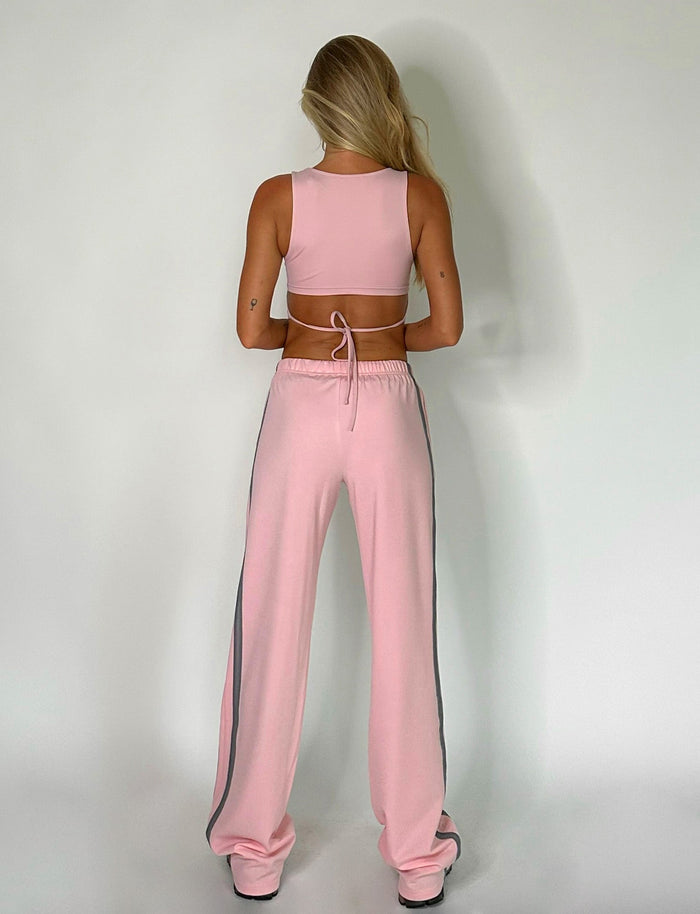 A person with long blonde hair wearing a TIGERMIST DARJA PANT in baby pink and a matching athletic crop top stands facing a white wall. The trousers feature faint gray stripes on each side. The person's hands are relaxed at their sides.