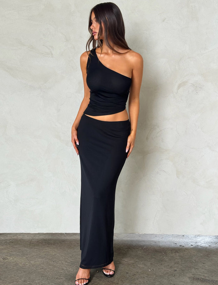 A woman with long, dark hair stands against a neutral background. She is wearing a one-shoulder, sleeveless black top from Tiger Mist, paired with the DEANNA MAXI SKIRT - BLACK from TIGERMIST and black open-toe heels. She has a relaxed and confident pose.
