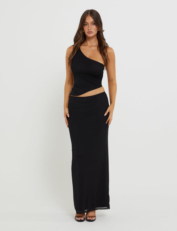 A woman with long brown hair is wearing a sleek black maxi skirt from TIGERMIST, known as the Deanna Maxi Skirt. She pairs it with a stylish one-shoulder top featuring a cut-out detail at the waist. She stands against a plain white background, looking directly at the camera and wearing black heeled sandals.