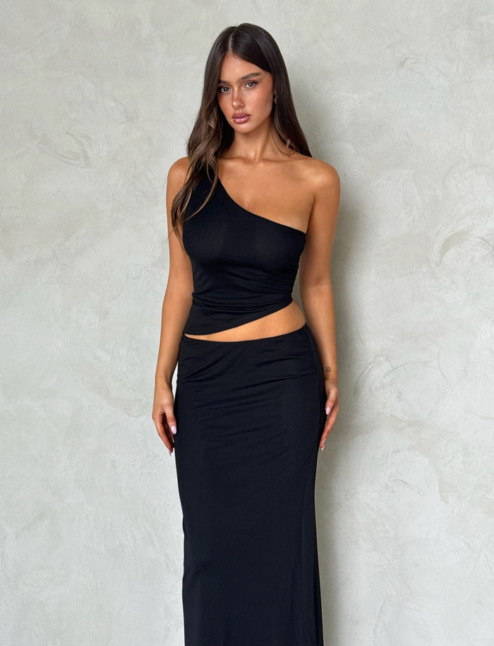 A woman stands against a textured, neutral wall. She is wearing the DEANNA MAXI SKIRT - BLACK and a black, one-shoulder top with a cutout at the waist from TIGERMIST. She has long, straight hair and a neutral expression, with her hands resting by her sides.