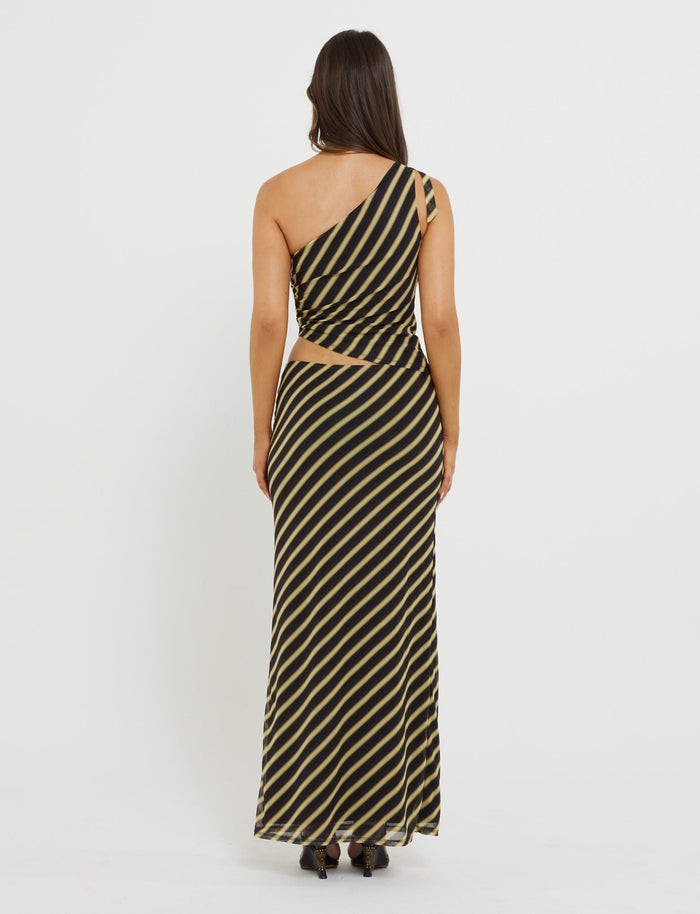 A person with long, dark hair stands facing away, wearing the DEANNA MAXI SKIRT - MULTI : STRIPE : BROWN STRIPE from TIGERMIST. The skirt features diagonal black and beige stripes and a cut-out detail on the right side at the waist. The person is also wearing black, open-toed heels.