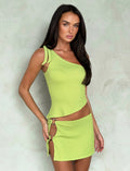 A woman wearing the DEANNA TOP - GREEN from TIGERMIST, a stylish lime green one-shoulder top, along with a matching mini skirt that features side cut-out designs and tie details, poses against a light-colored textured background. She has long, dark hair and is looking directly at the camera.