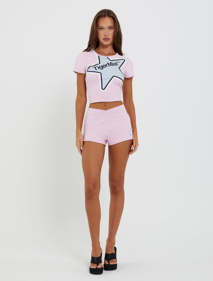 A woman stands against a white background wearing a pink crop top featuring a star design and the text "TIGERMIST." She pairs it with the DELANEY SHORT - PINK, which has a bodycon fit, low-rise waist, and completes her look with black heels.