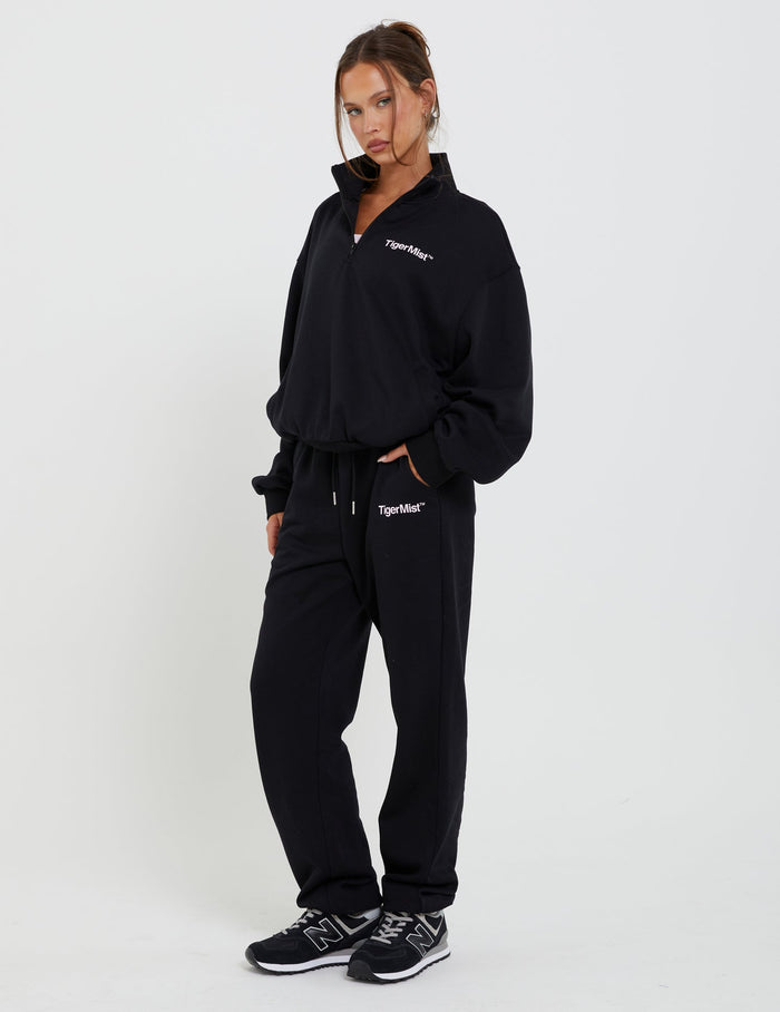 A person stands wearing the DELFINA JUMPER - BLACK and matching black sweatpants, both emblazoned with "TIGERMIST." With hands in pockets and black sneakers completing the look, they pose confidently against a plain white background.