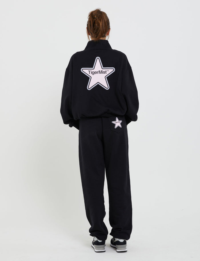 A person stands against a white backdrop, dressed in a black tracksuit adorned with a prominent white star and the "TIGERMIST" logo on the back. The coordinating black pants, named DELFINA PANT - BLACK, showcase a small white star near the waistband. The individual's hair is tied up and they are wearing black and white sneakers.