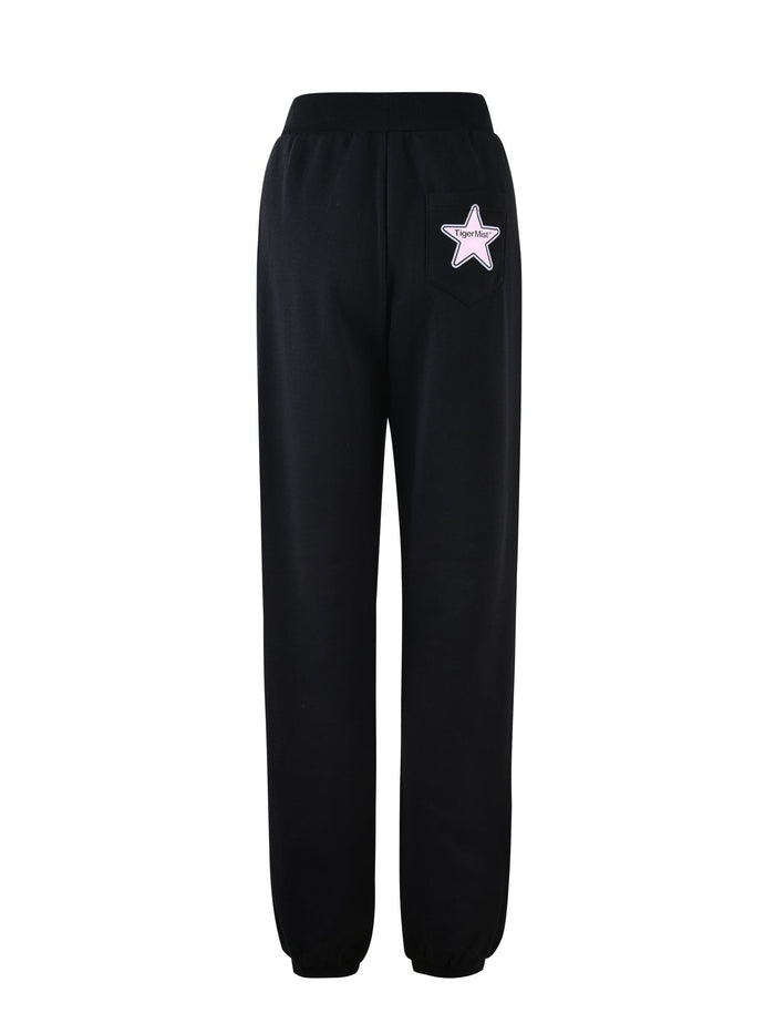 The DELFINA PANT - BLACK by TIGERMIST is a pair of black jogger pants with elastic cuffs. The back right pocket features a pink star patch with the text "smile" written inside the star. These joggers have a relaxed fit and appear to be made of comfortable, soft material.