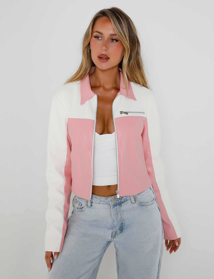 A woman with long blonde hair stands against a plain white background. Clad in a stylish TIGERMIST DELIA JACKET - PINK : PINK/WHITE with a front zipper over a white top, she pairs it with light blue high-waisted jeans. Her expression is neutral as she gazes slightly to the side.