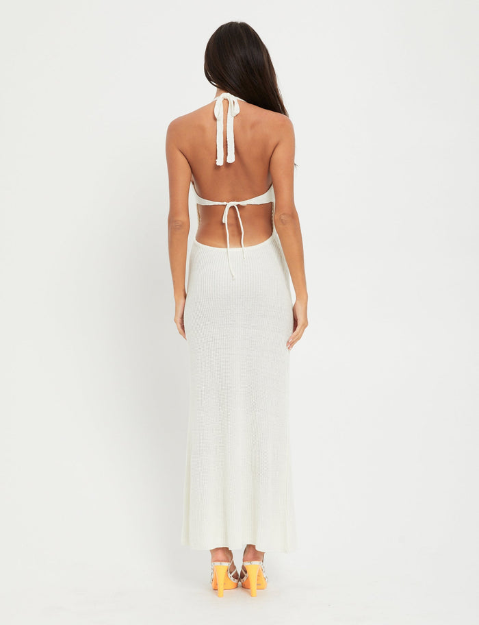 A woman with long dark hair is seen from the back wearing the DELTA DRESS - WHITE, a backless halter neck dress by TIGERMIST that ties at the neck and lower back. She is also wearing high heel sandals with neon orange soles. The background is plain white.