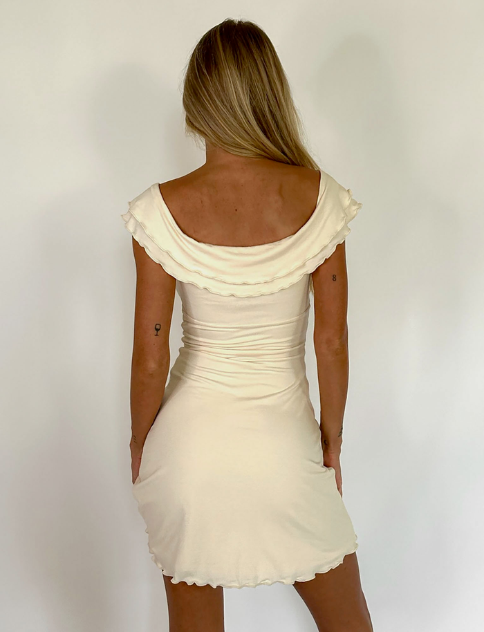 A woman with long blonde hair faces away, wearing the off-the-shoulder, fitted TIGERMIST DENALI MINI DRESS in white bone with ruffled edges and buttery jersey fabric, set against a plain white background.