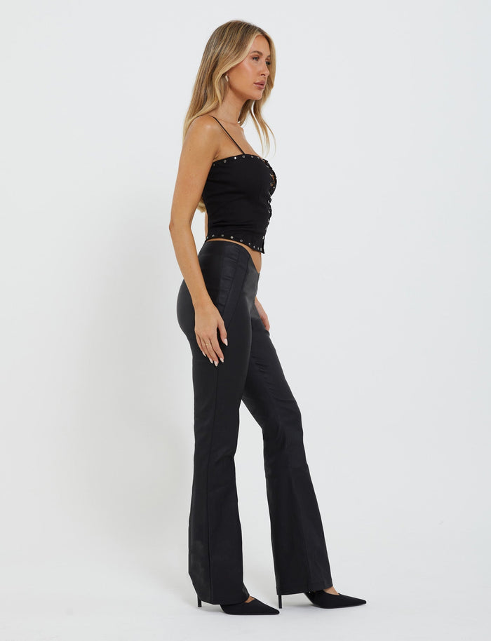 A woman with long blonde hair, dressed in a sleek, black strapless top and the high-waisted DOPHA PANT - BLACK from TIGERMIST, stands in profile against a plain white background. She completes her stylish look with black pointed-toe heels, gazing forward with a neutral expression.